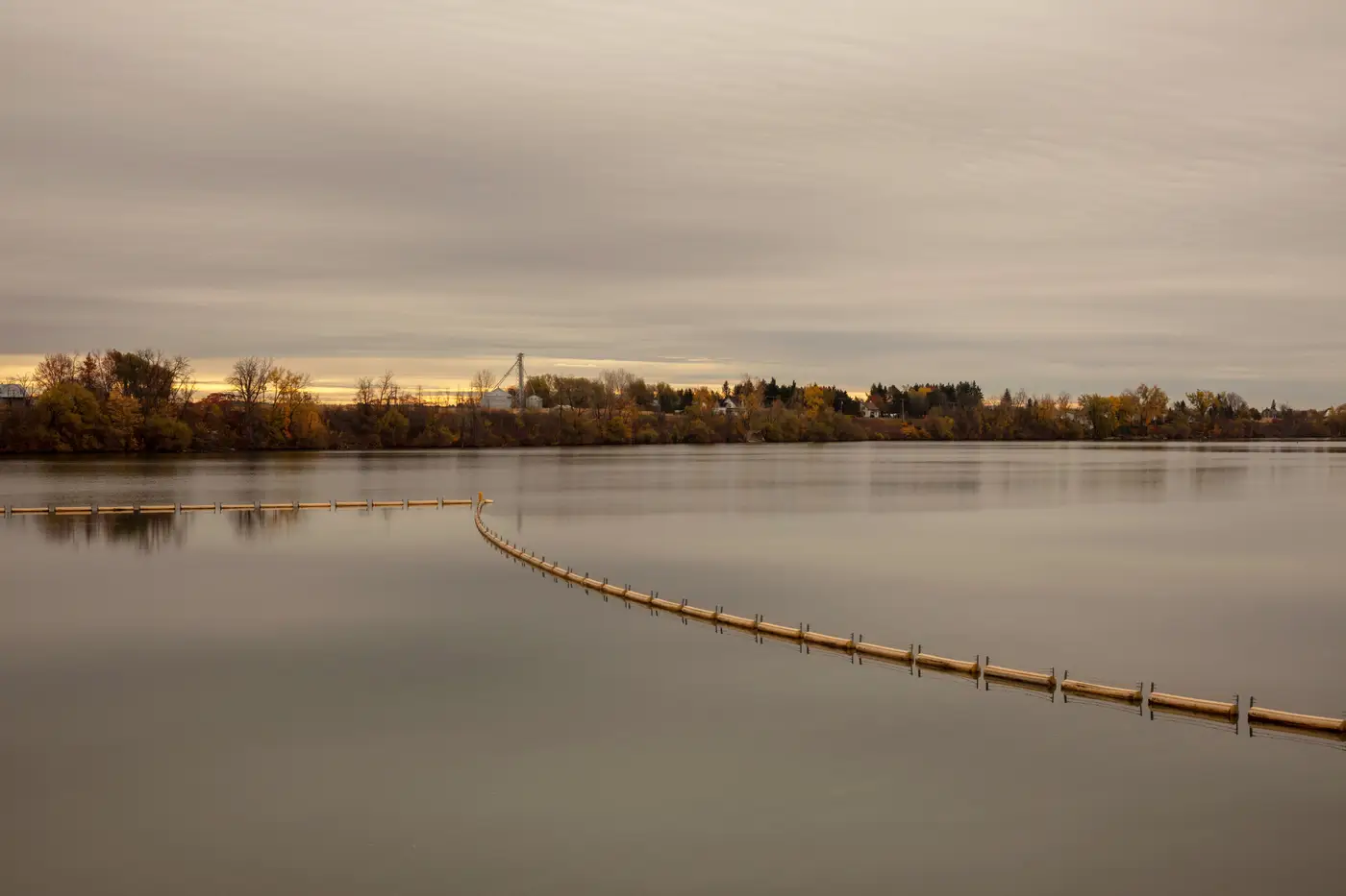 A floating boom strung across calm water, curving from the near bank toward the far shore.